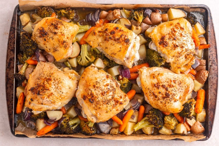 Roasted chicken thighs topped with garlic butter sauce arranged atop a bed of vegetables on a sheet pan.