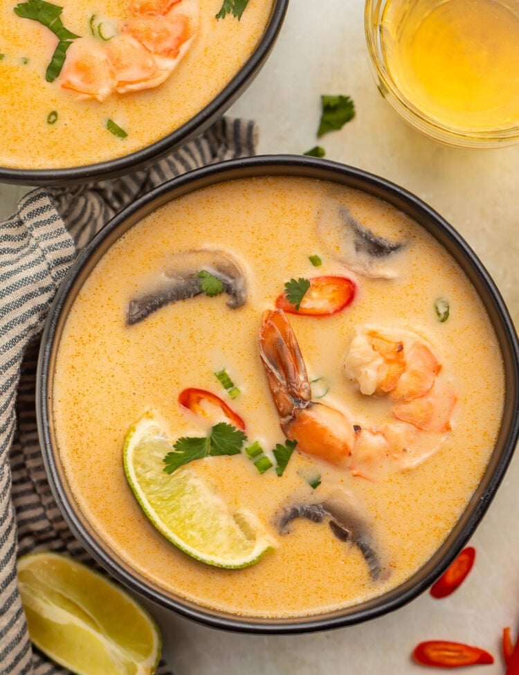 An overhead photo of a black soup bowl holding tom kha goong (Thai coconut shrimp soup) with lime wedges, shrimp, and aromatics floating in the broth.