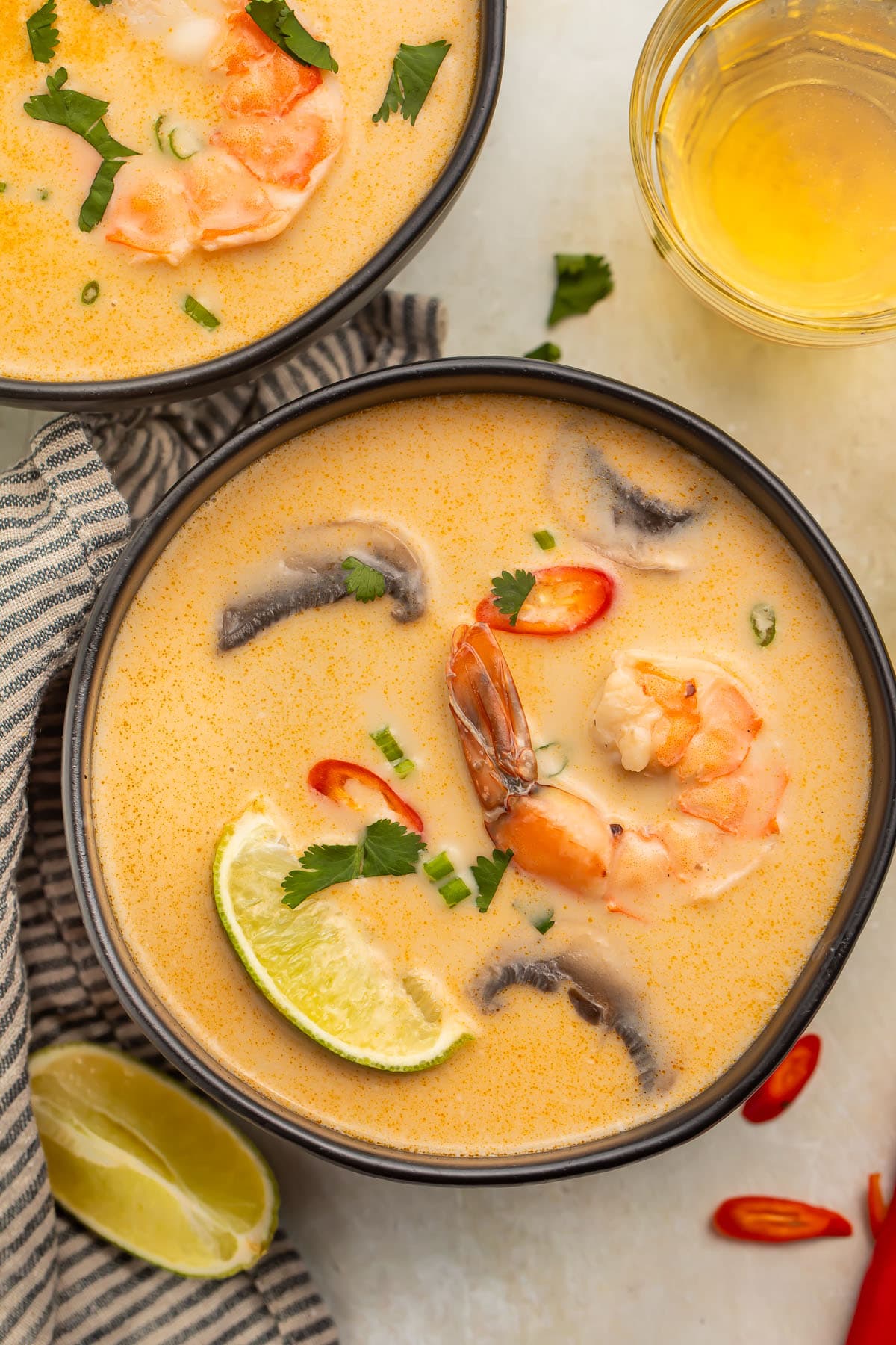 An overhead photo of a black soup bowl holding tom kha goong (Thai coconut shrimp soup) with lime wedges, shrimp, and aromatics floating in the broth.
