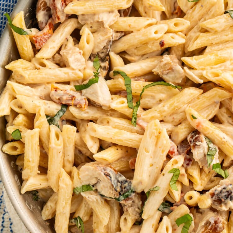 A large silver skillet of penne pasta in a sun-dried tomato cream sauce with chicken and basil.