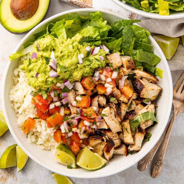 A large vibrant chicken burrito bowl with cauliflower rice, diced chicken, fresh pico de gallo, guacamole, and lime wedges.