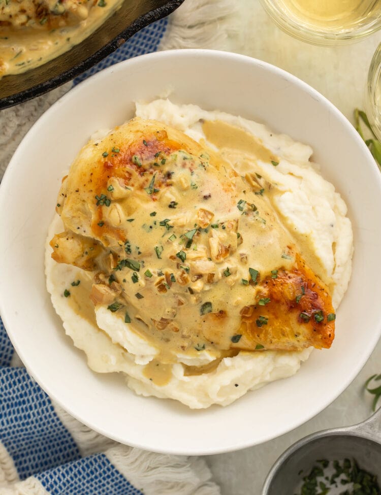 Creamy dijon chicken breast placed on top of a bed of mashed potatoes in a large white bowl on a table with a blue and white stripe dish towel.
