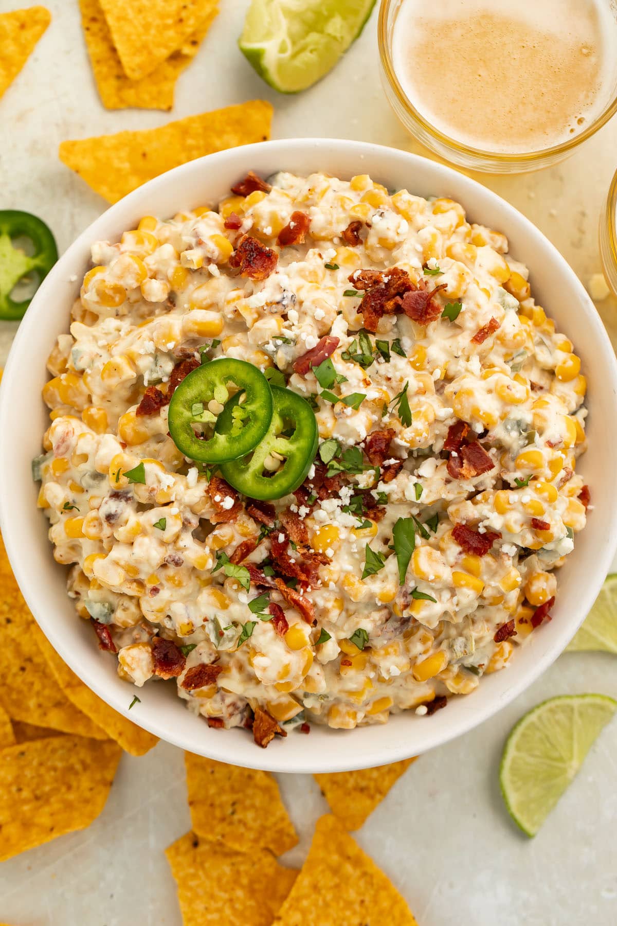 A top-down view of a large white bowl of jalapeño corn dip surrounded by tortilla chips on a table.