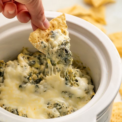 A woman using a tortilla chip to scoop Crockpot spinach artichoke dip out of a slow cooker.