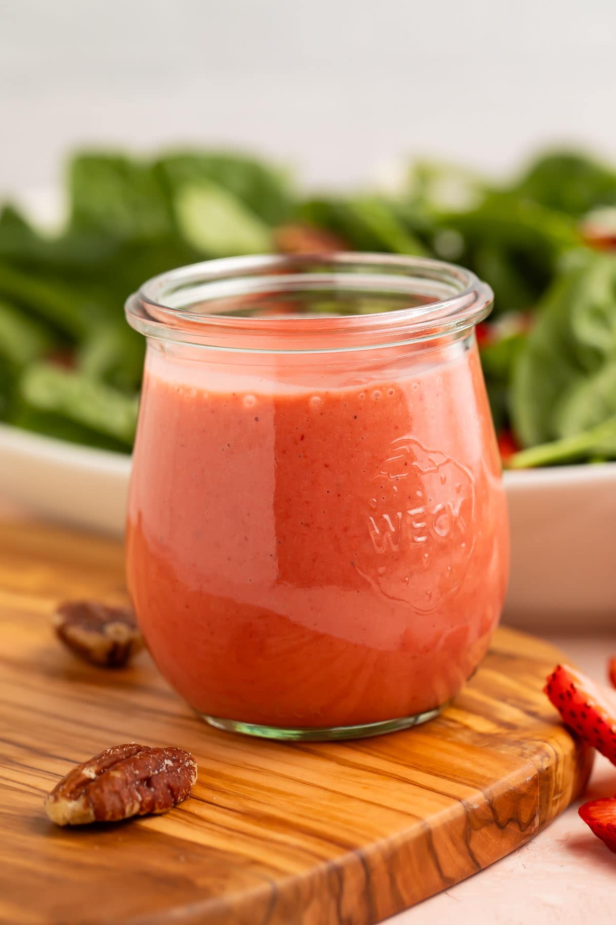 A jar of creamy pale red strawberry vinaigrette in front of a leafy spinach salad.