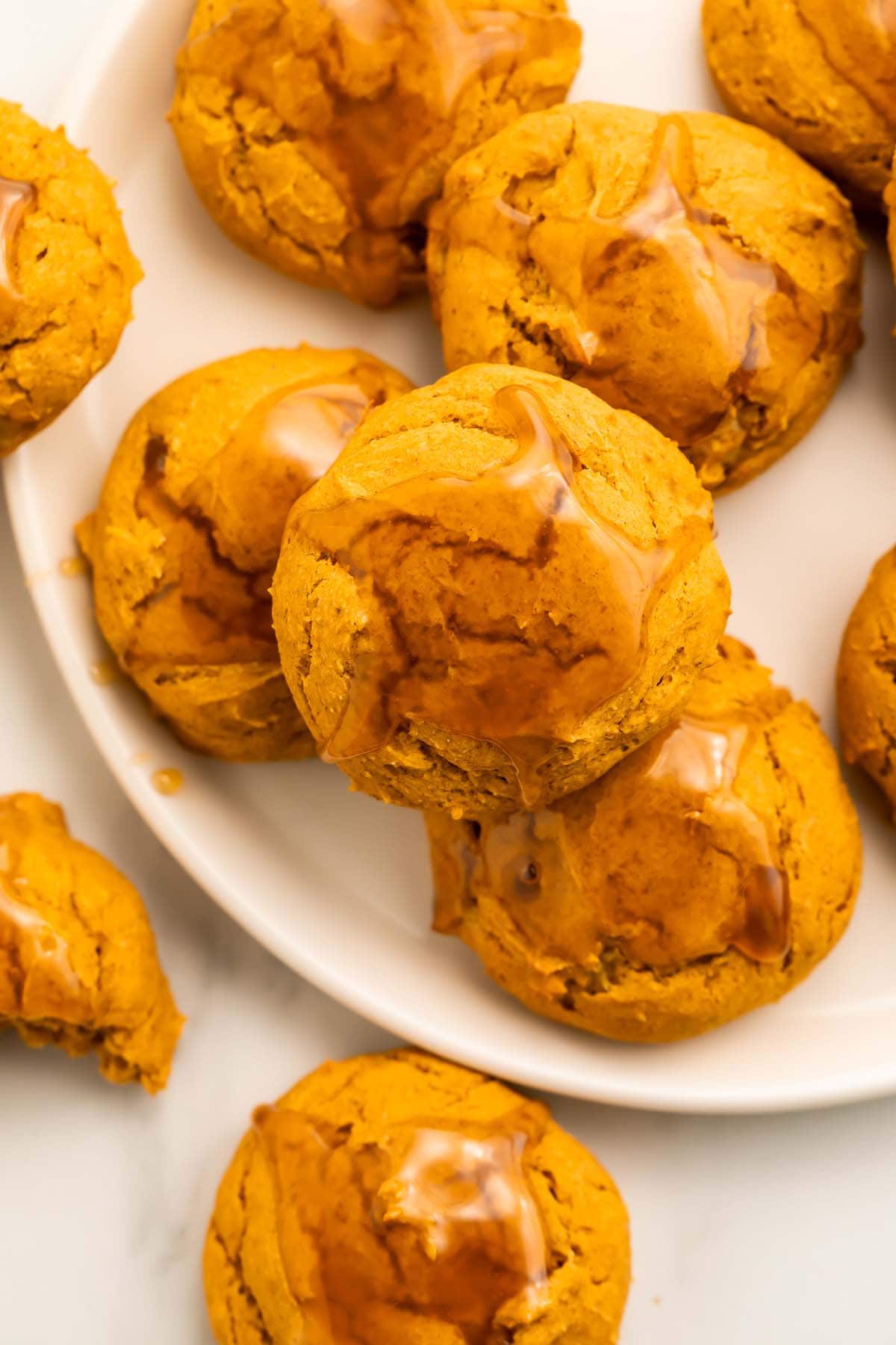 Pumpkin cookies topped with a light maple glaze arranged on and around a round white plate on a table.