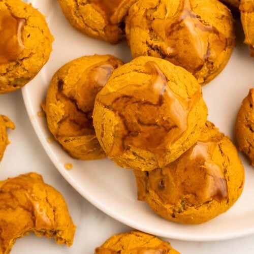 Pumpkin cookies topped with a light maple glaze arranged on and around a round white plate on a table.