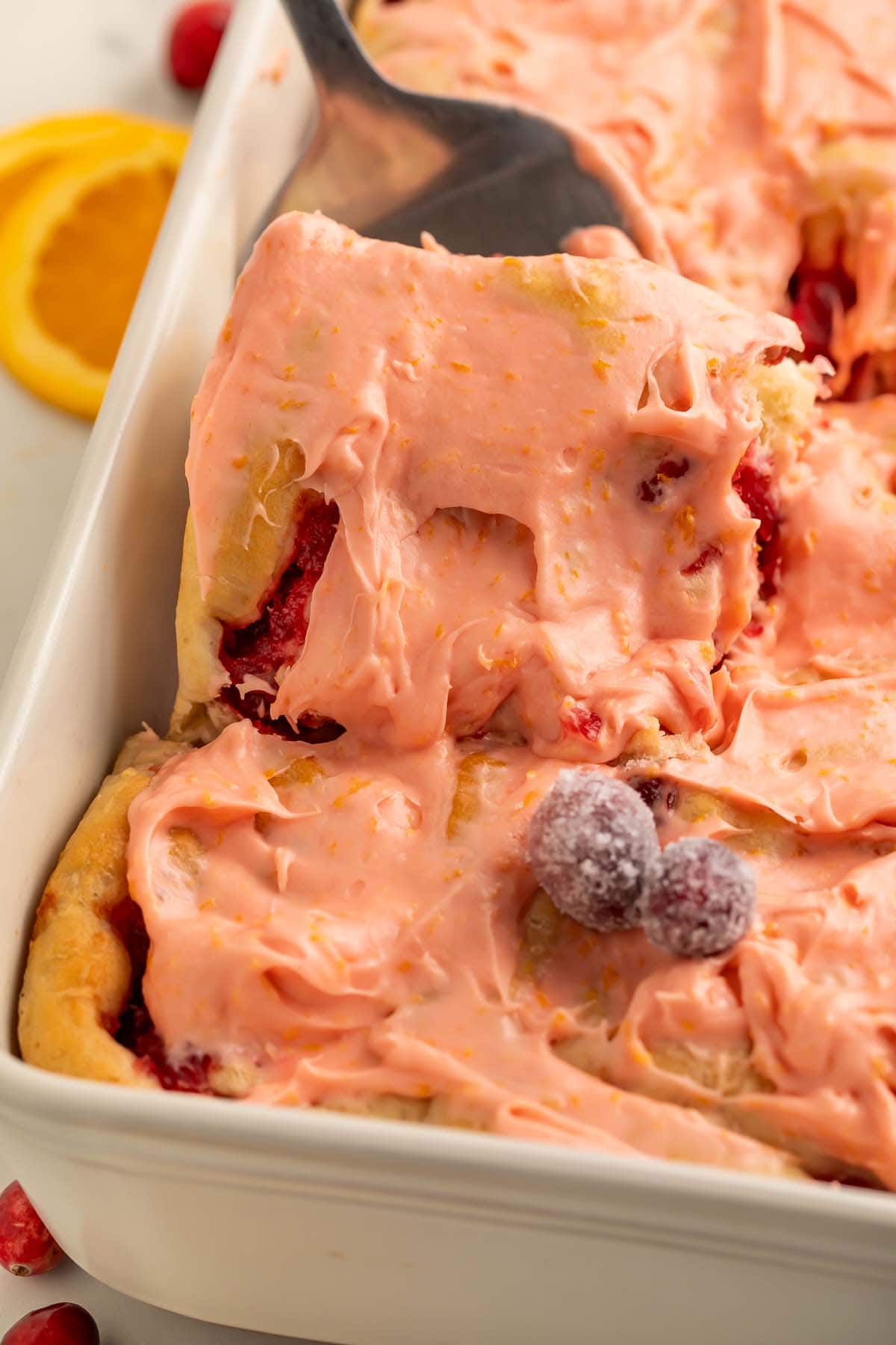 A spatula lifts a pink-frosted cranberry cinnamon roll out of a pan of cinnamon rolls.