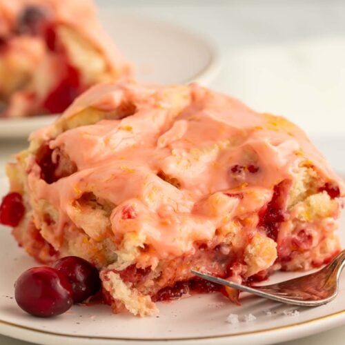 A cranberry orange cinnamon roll resting on a small plate with 2 loose cranberries.