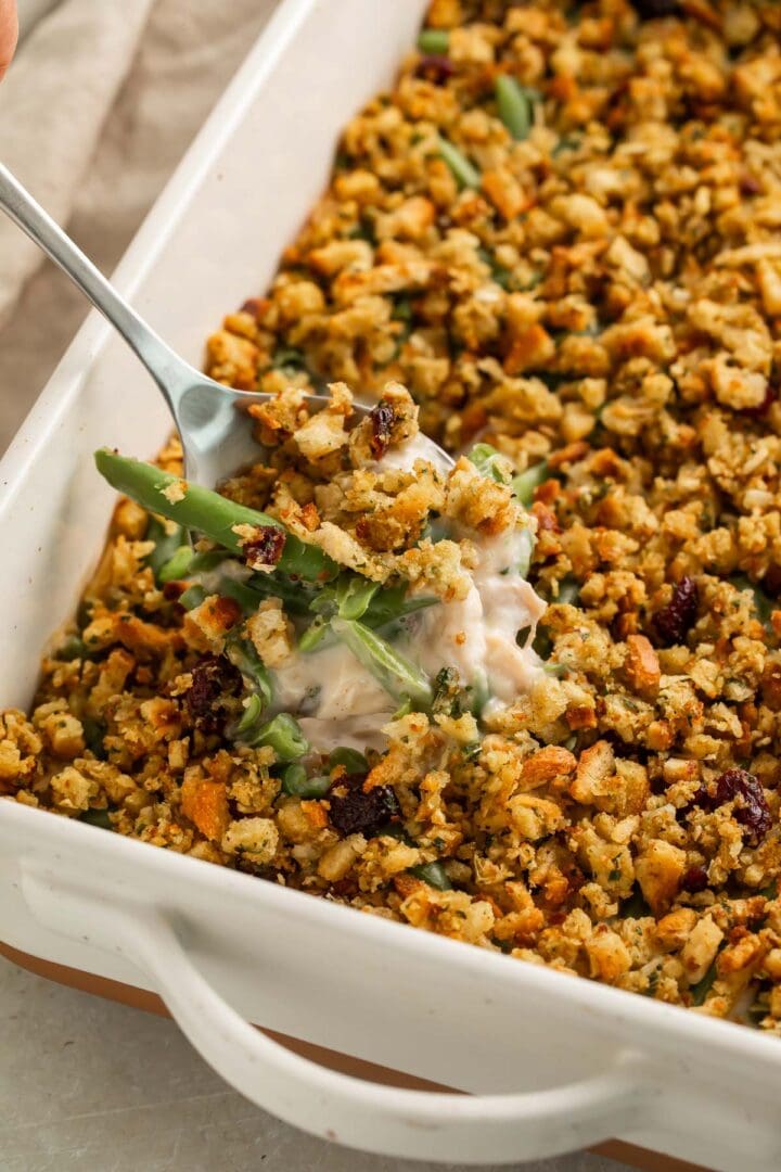 A scoop of creamy chicken and stuffing casserole being lifted out of a casserole dish, showing the creamy sauce and green beans inside.