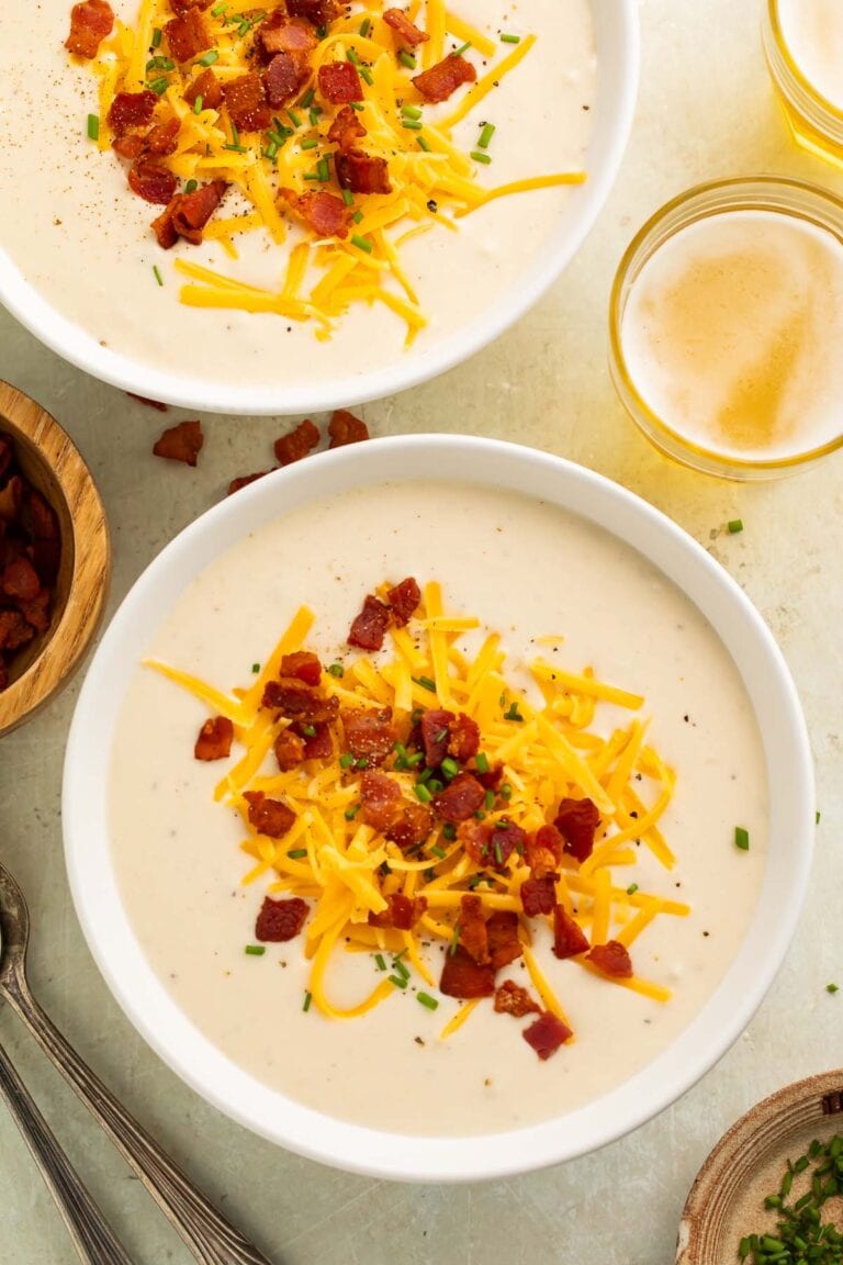 Creamy mashed potato soup in white bowls topped with shredded cheddar cheese, crispy bacon bits, and fresh green onions