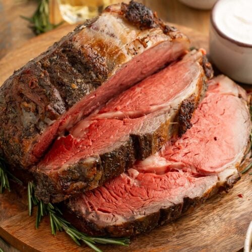 A thickly-carved reverse sear prime rib on a wooden cutting board next to prepared horseradish sauce and au jus.