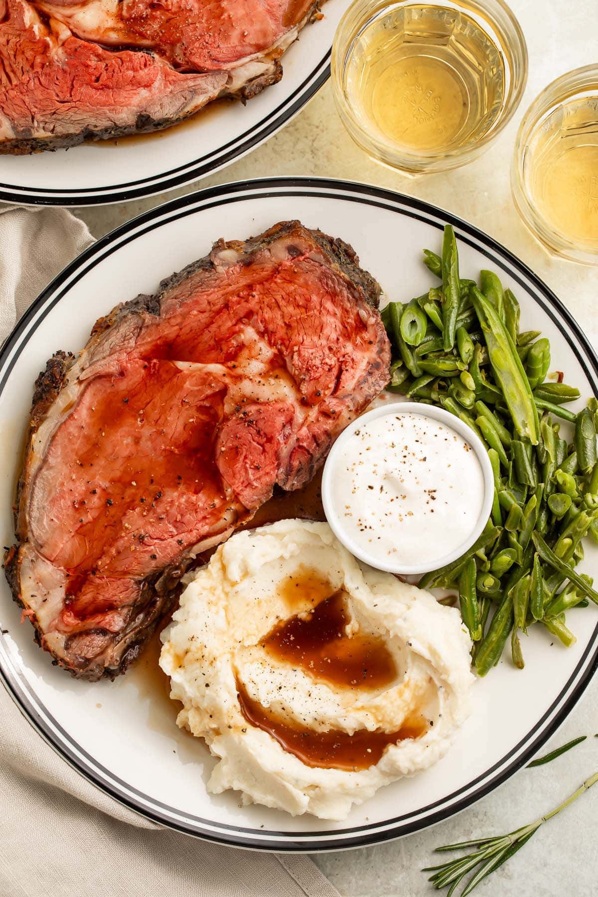 Overhead photo of a plate round white plate with a dark border. On the plate rests a slice of reverse sear prime rib, a scoop of mashed potatoes and gravy, green beans, and horseradish sauce.