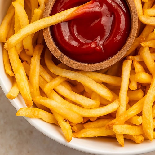 Golden air fryer frozen french fries on a white plate with ketchup in a wooden bowl.