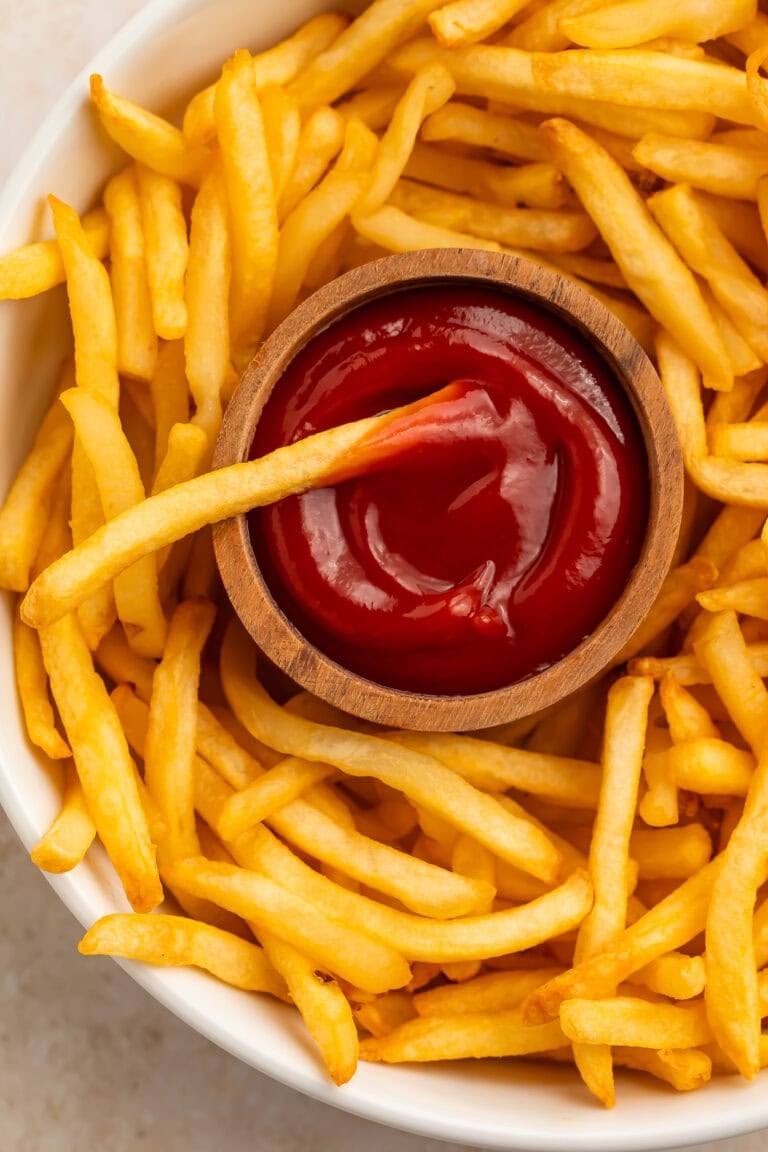 Golden air fryer frozen french fries on a white plate with ketchup in a wooden bowl.