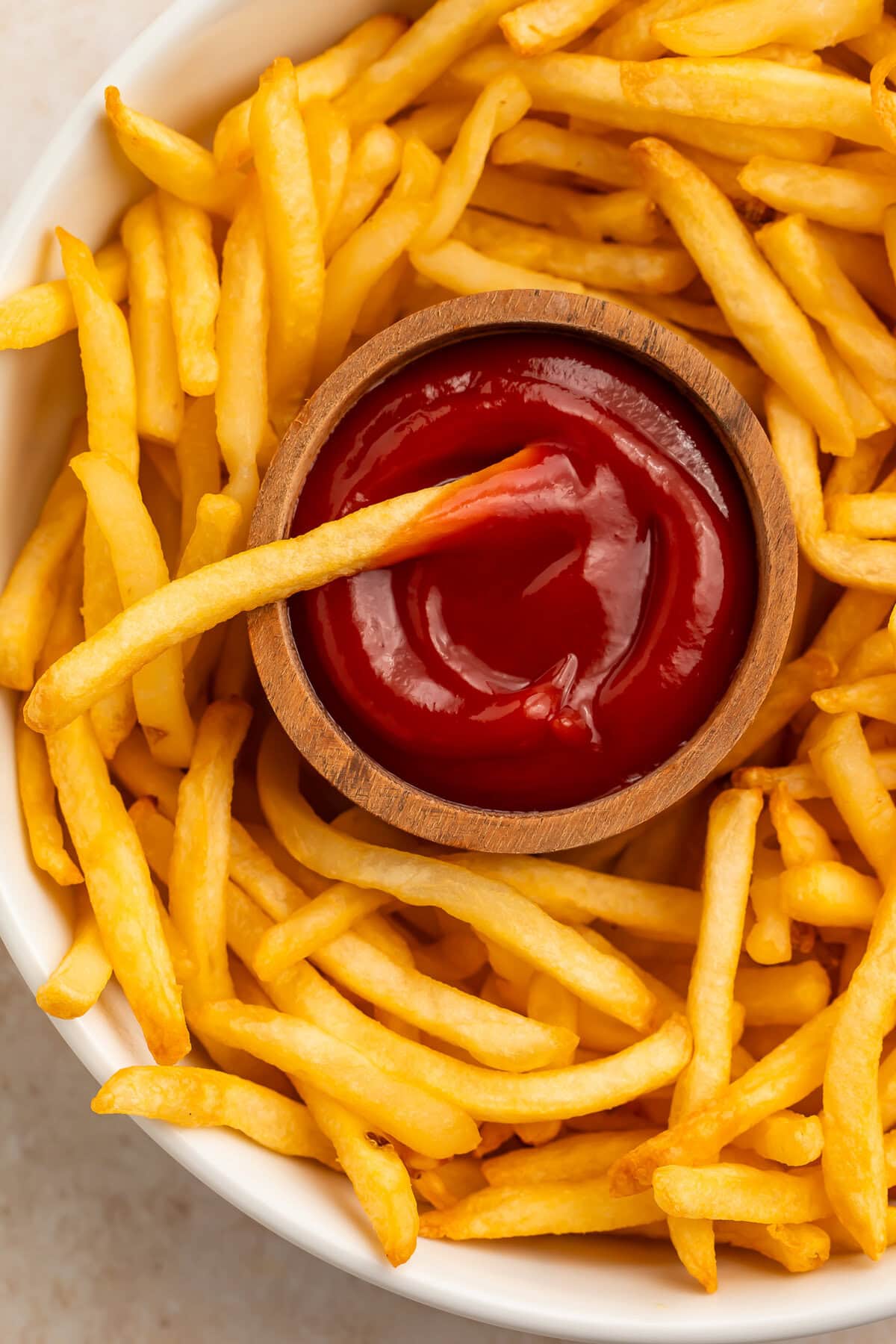Golden air fryer frozen french fries on a white plate with ketchup in a wooden bowl.