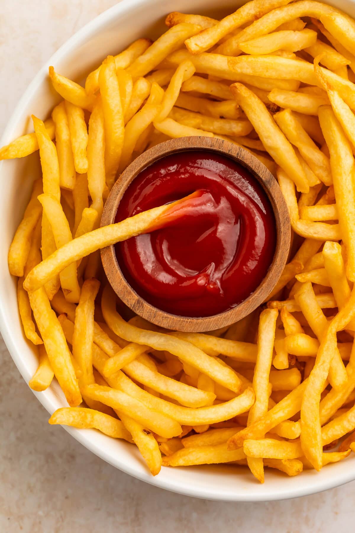 Crispy golden french fries surrounding a wooden bowl of ketchup on a white plate.