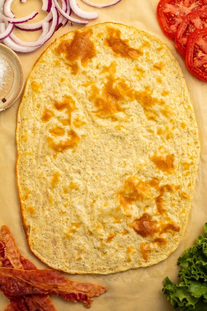 An unfolded cottage cheese flatbread on a sheet of parchment paper surrounded by sandwich ingredients.