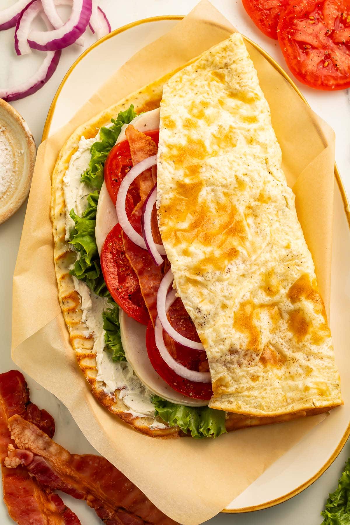 A flatbread sandwich of ham, bacon, tomato, lettuce, and onion on a cottage cheese flatbread.