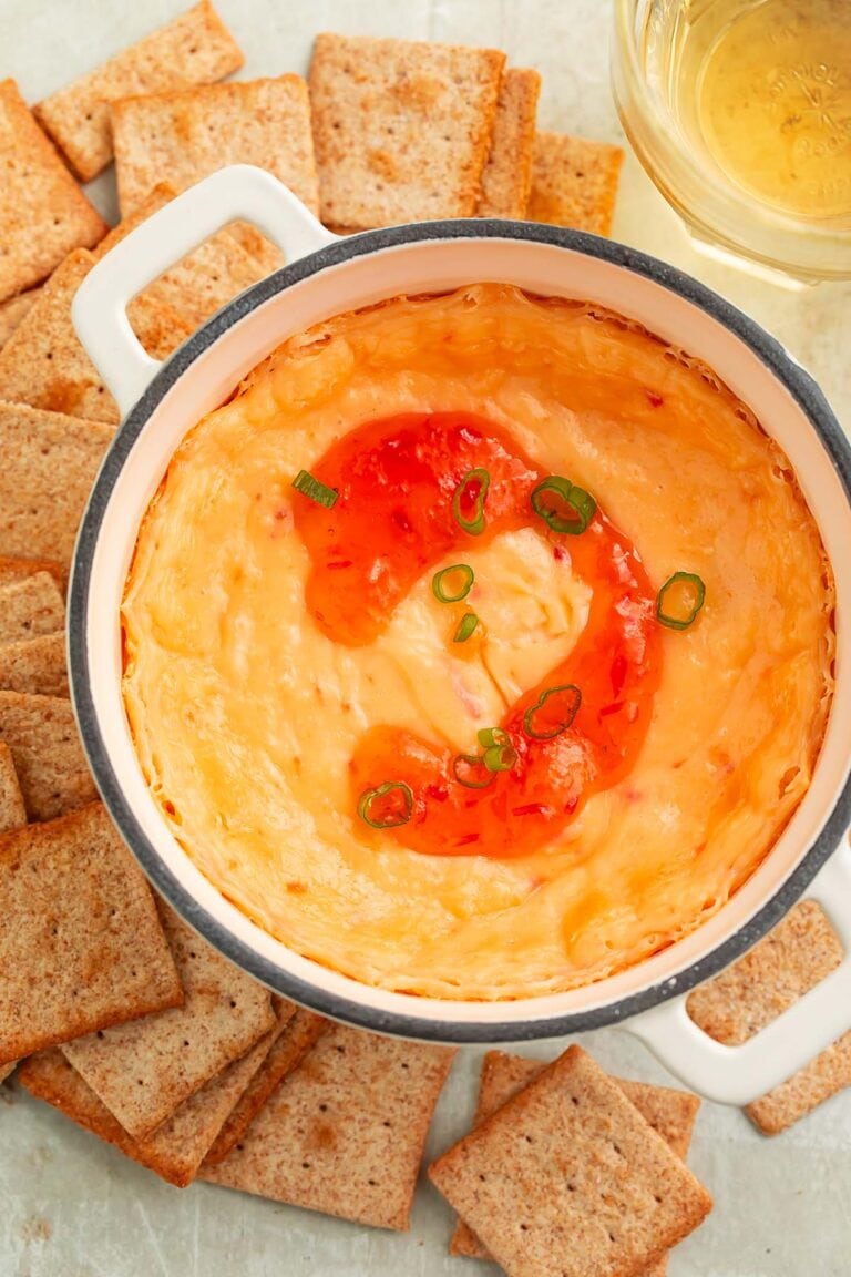 A high-sided bowl of cream cheese pepper jelly dip topped with a swirl of red pepper jelly, surrounded by square wheat crackers.