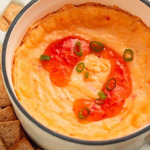 A high-sided bowl of cream cheese pepper jelly dip topped with a swirl of red pepper jelly, surrounded by square wheat crackers.