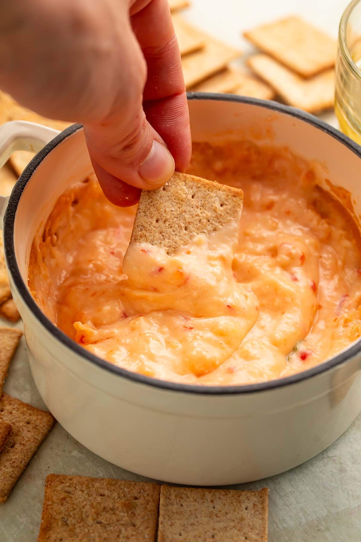 A white woman's hand holding a square cracker that she's dipping into a high-sided bowl of cream cheese red pepper jelly dip.