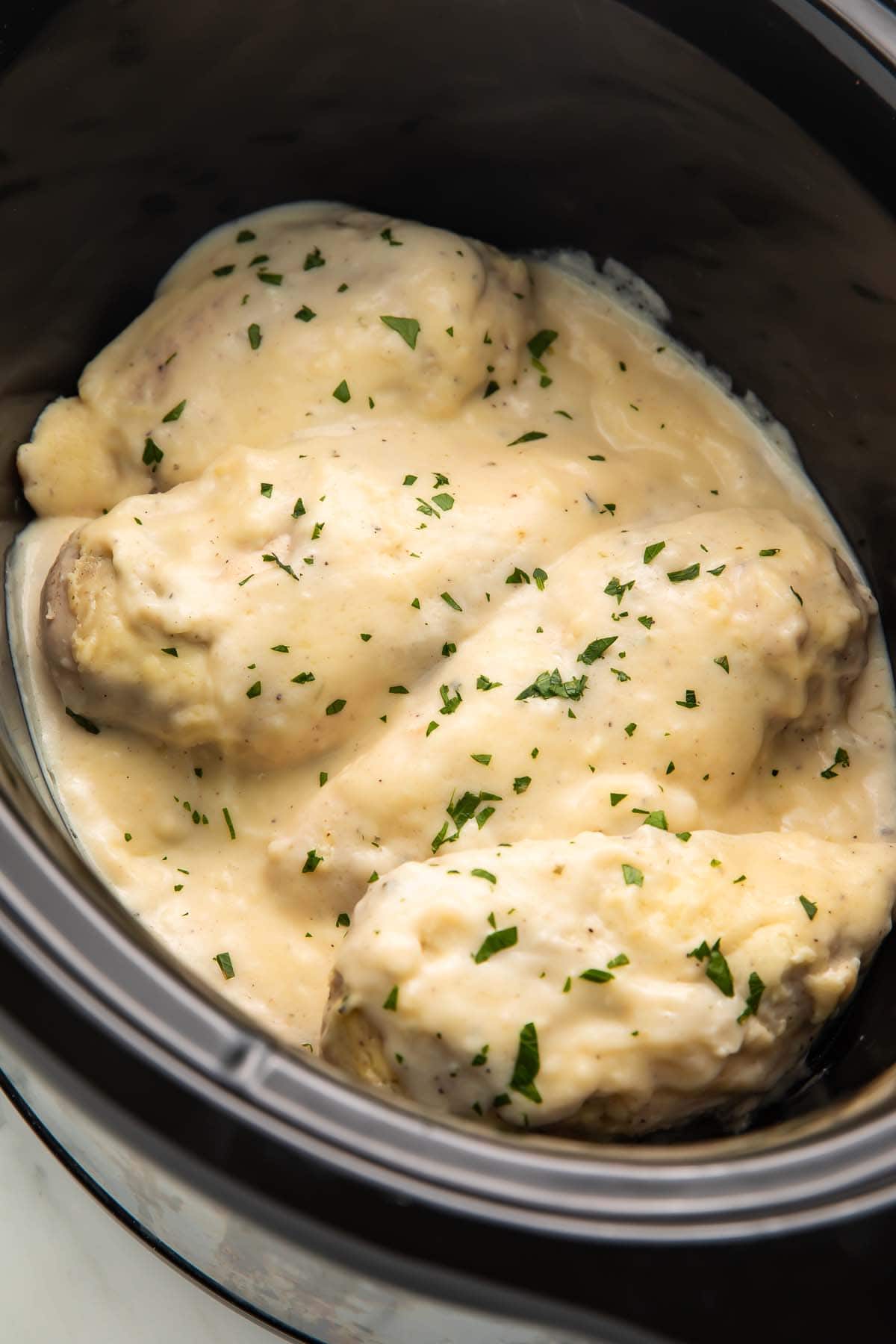 Four medium chicken breasts topped with creamy Boursin sauce and chopped chives in a black ceramic Crockpot insert.