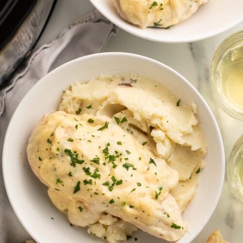 A white bowl containing a bed of mashed potatoes topped with a creamy Crockpot Boursin chicken breast on a table.