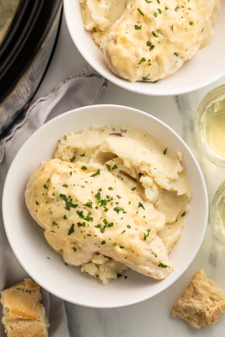 A white bowl containing a bed of mashed potatoes topped with a creamy Crockpot Boursin chicken breast on a table.