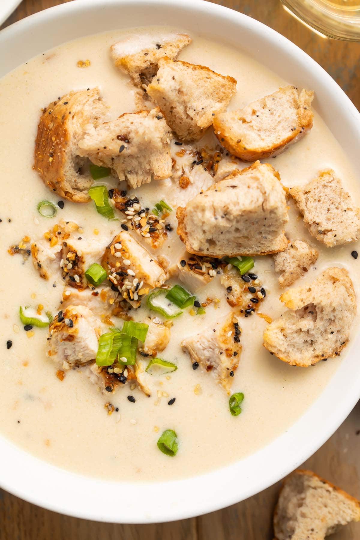 A close-up look at a large white bowl filled with a cream-based chicken soup, topped with everything bagel croutons and sliced green onions.