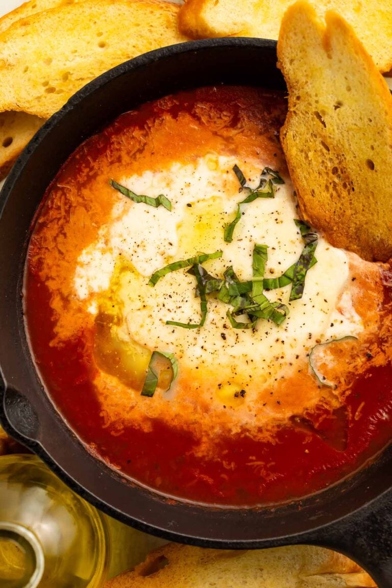 A large black bowl holding rich red marinara and truffle baked burrata garnished with thinly sliced fresh basil and a piece of crostini.