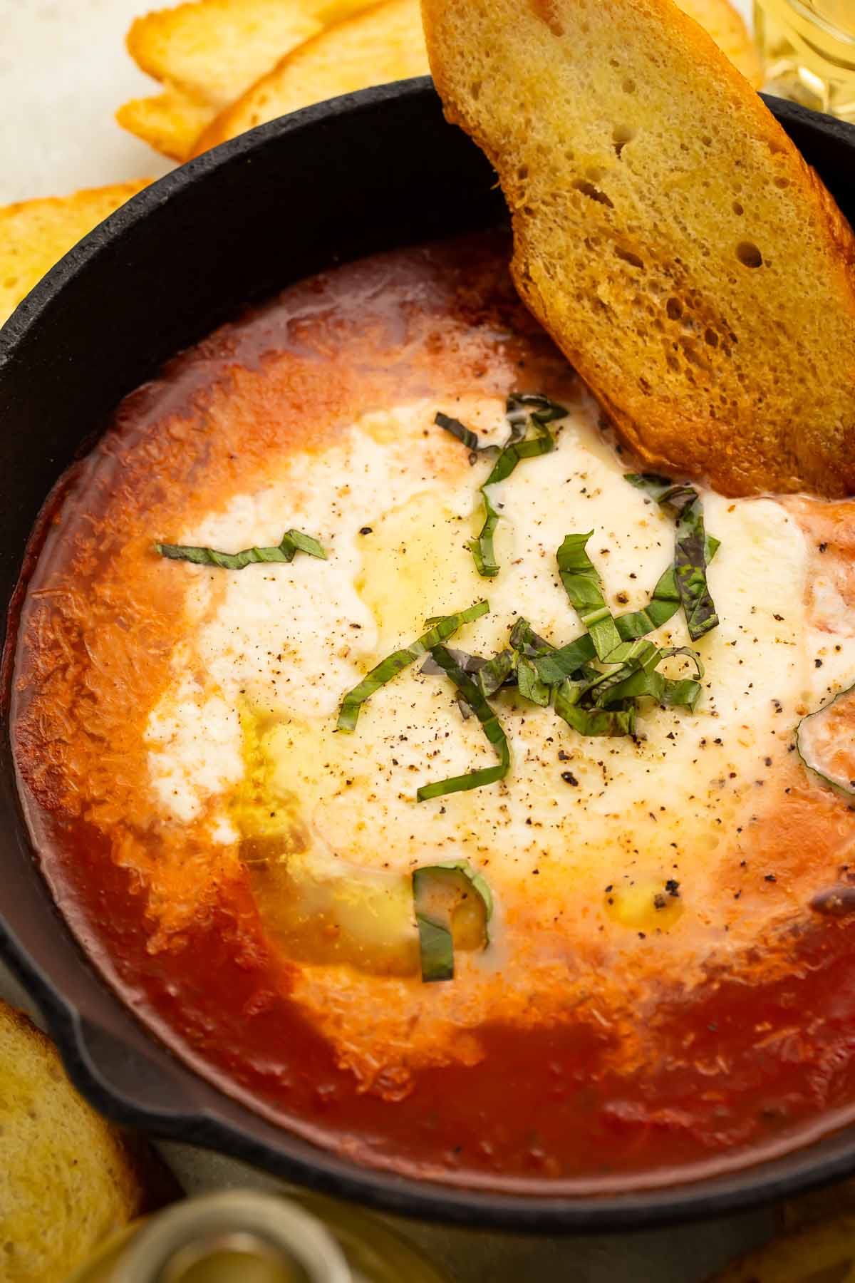 A large black bowl holding rich red marinara and truffle baked burrata garnished with thinly sliced fresh basil and a piece of crostini.