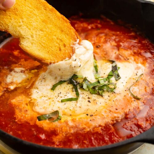 A piece of crostini scooping marinara and truffle baked burrata out of a black bowl.