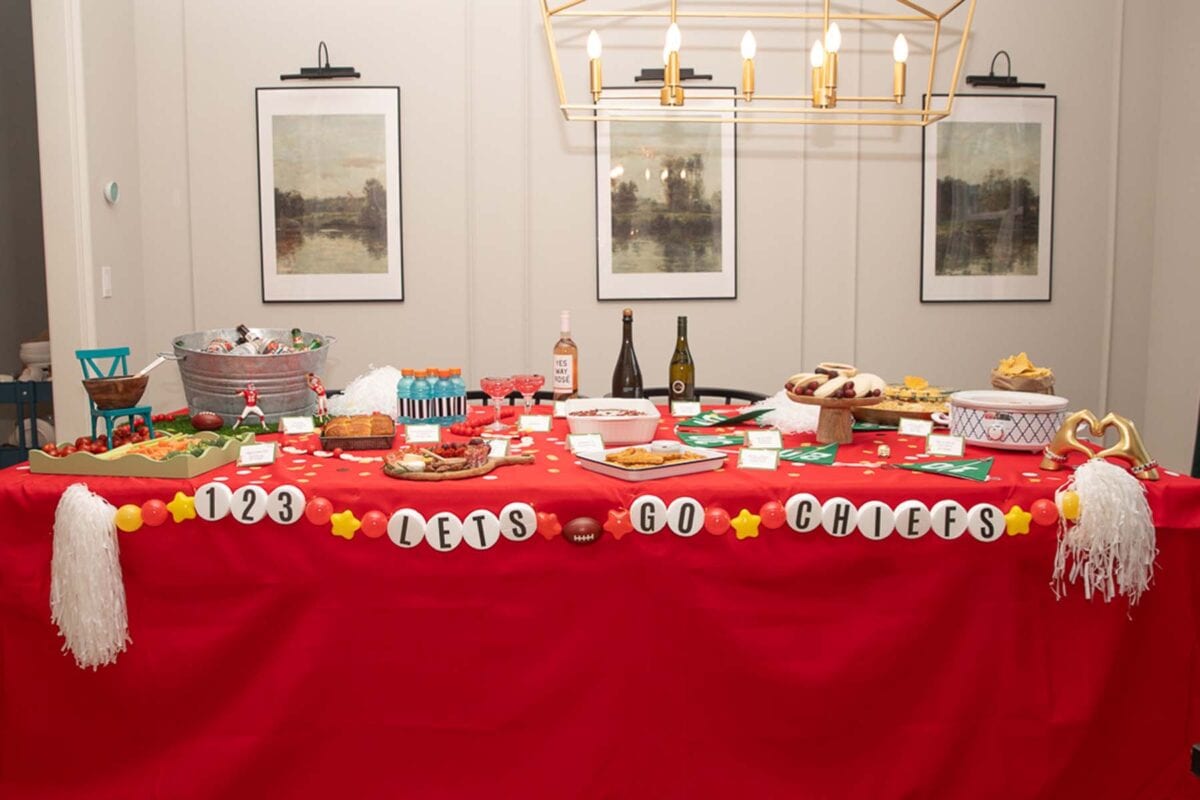 Dining room table, fully decorated with Taylor Swift Super Bowl decorations and food. A bracelet garland across the front of the table reads "1, 2, 3, Let's Go Chiefs".