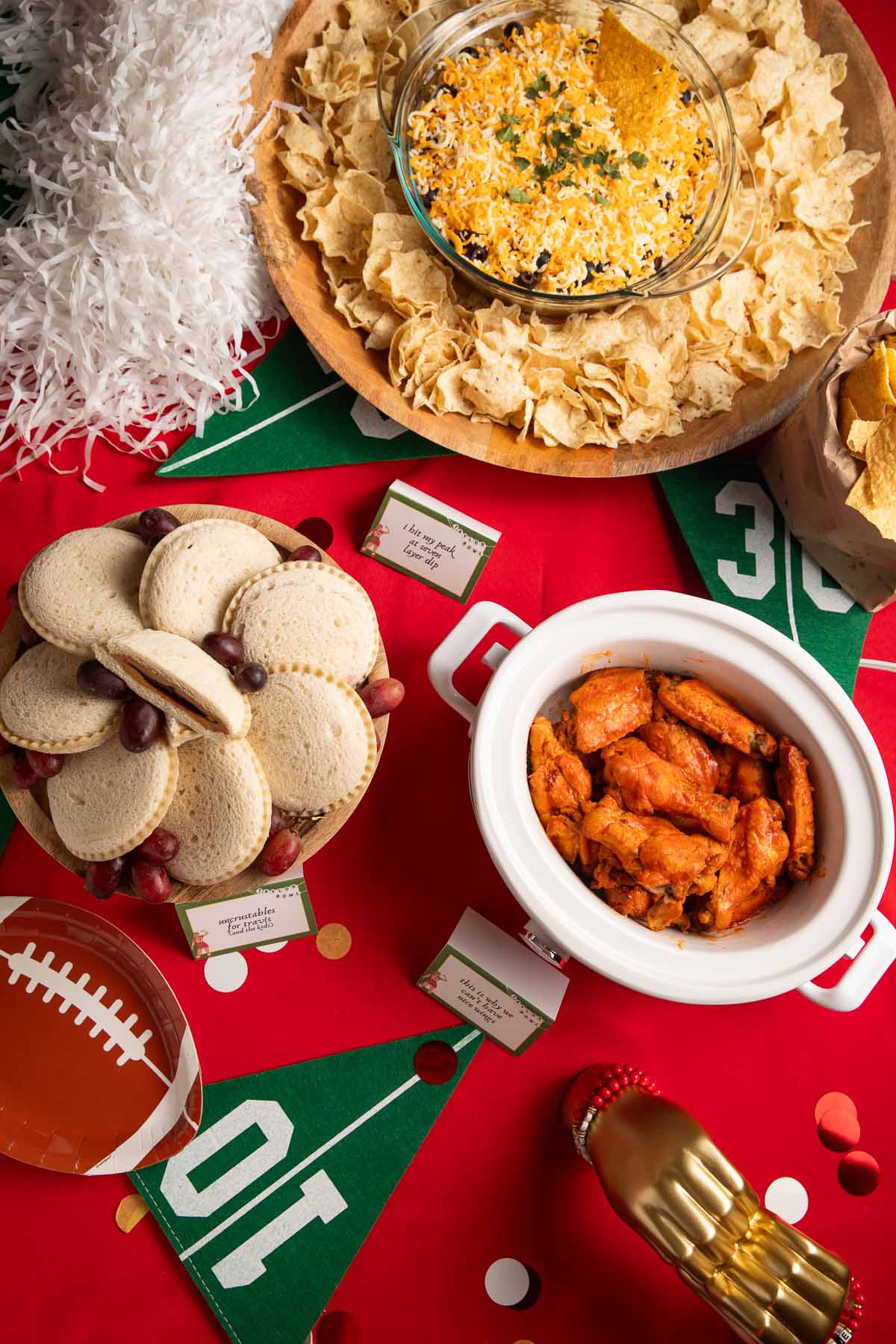 A table set for a Taylor Swift Super Bowl party, with football decorations, a red tablecloth, and gold heart hands placed among Crockpot chicken wings, Uncrustable sandwiches, and a 7 layer dip.