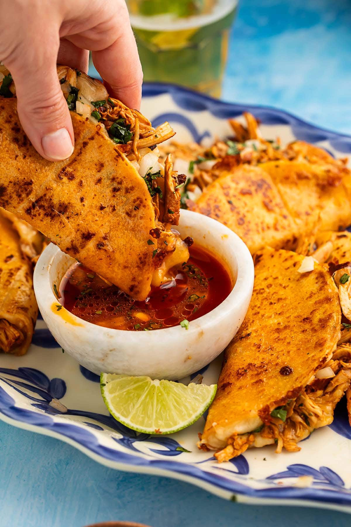 A white woman's hand holds a chicken birria taco, dipping one end of the taco into a ramekin of red salsa.