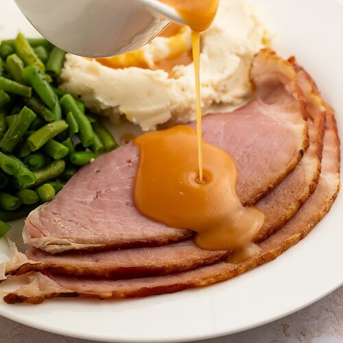 Creamy, richly brown ham gravy being poured from a gravy boat onto slices of ham plated with green beans and mashed potatoes.