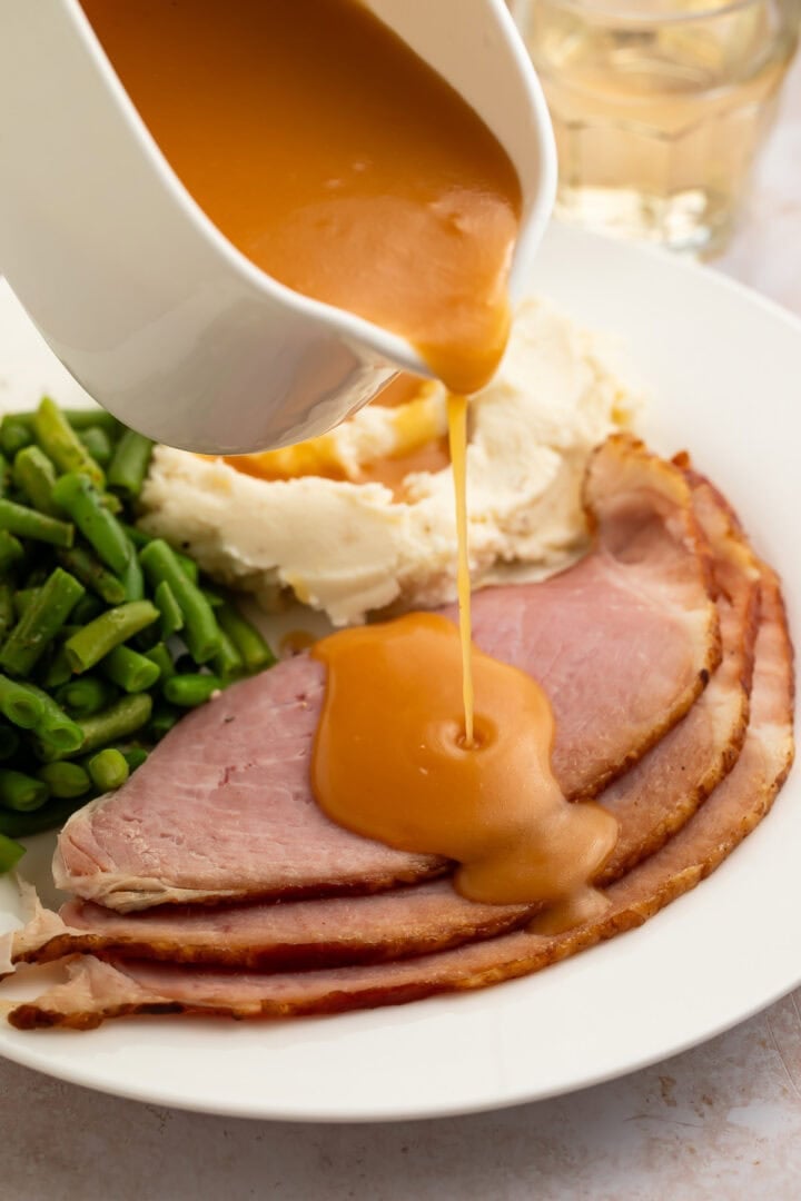 Creamy, richly brown ham gravy being poured from a gravy boat onto slices of ham plated with green beans and mashed potatoes.