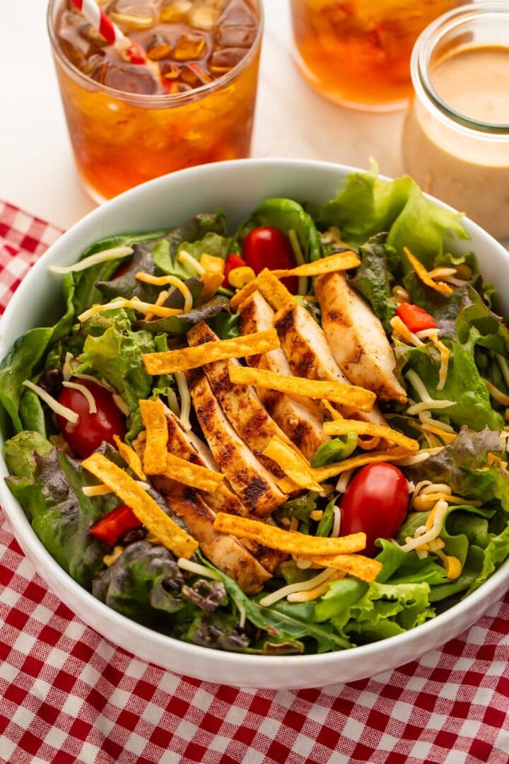 A shallow white bowl holding a copycat Chick-fil-A southwest salad with slices of grilled chicken, crispy tortilla strips, and red cherry tomatoes.