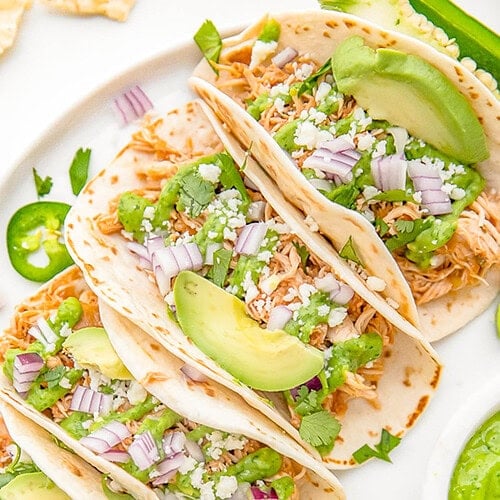 Crockpot chicken tacos arranged on a white plate. The tacos are topped with diced red onion, slices of avocado, and crumbled cheese.
