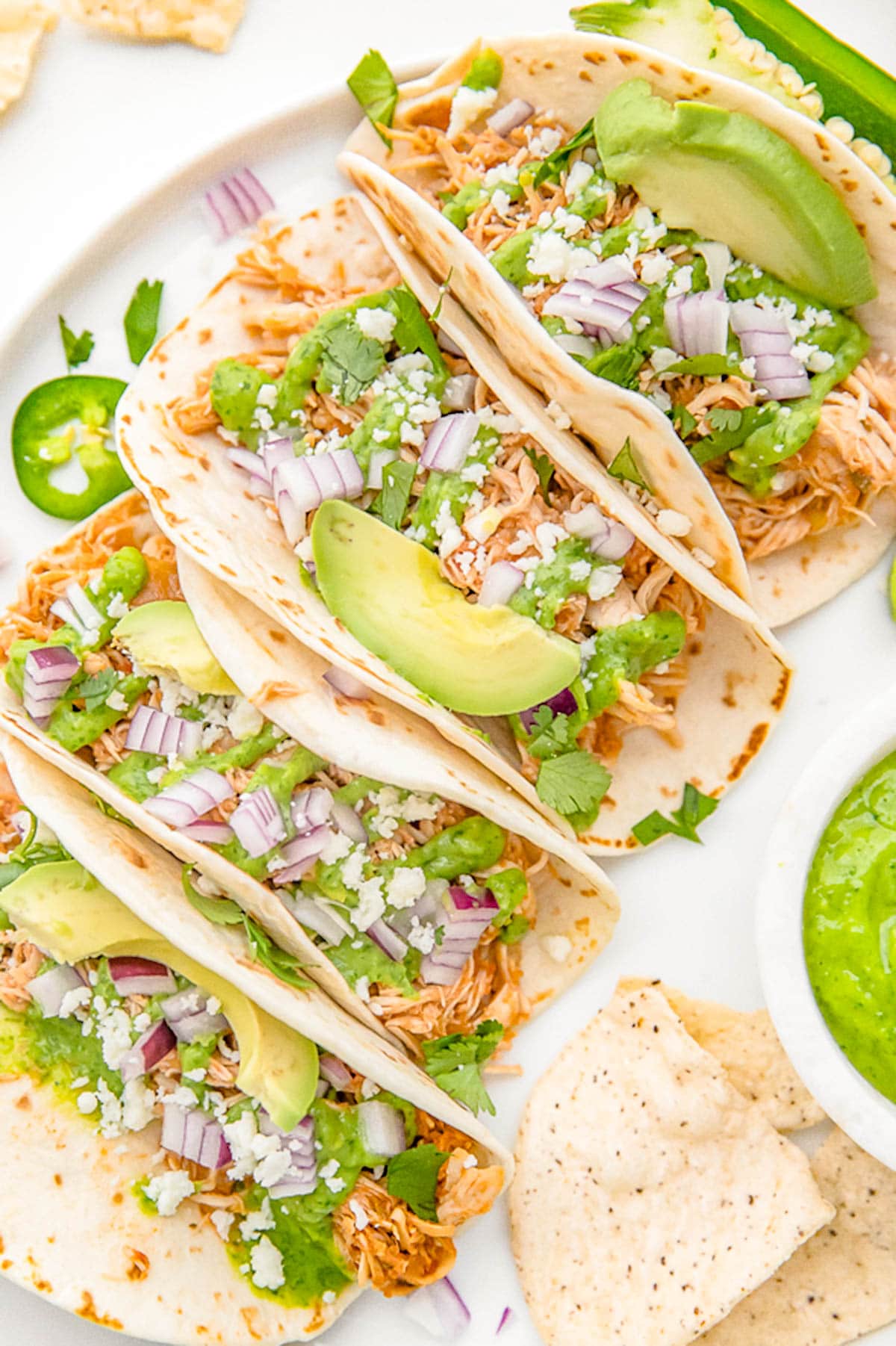 Crockpot chicken tacos arranged on a white plate. The tacos are topped with diced red onion, slices of avocado, and crumbled cheese.