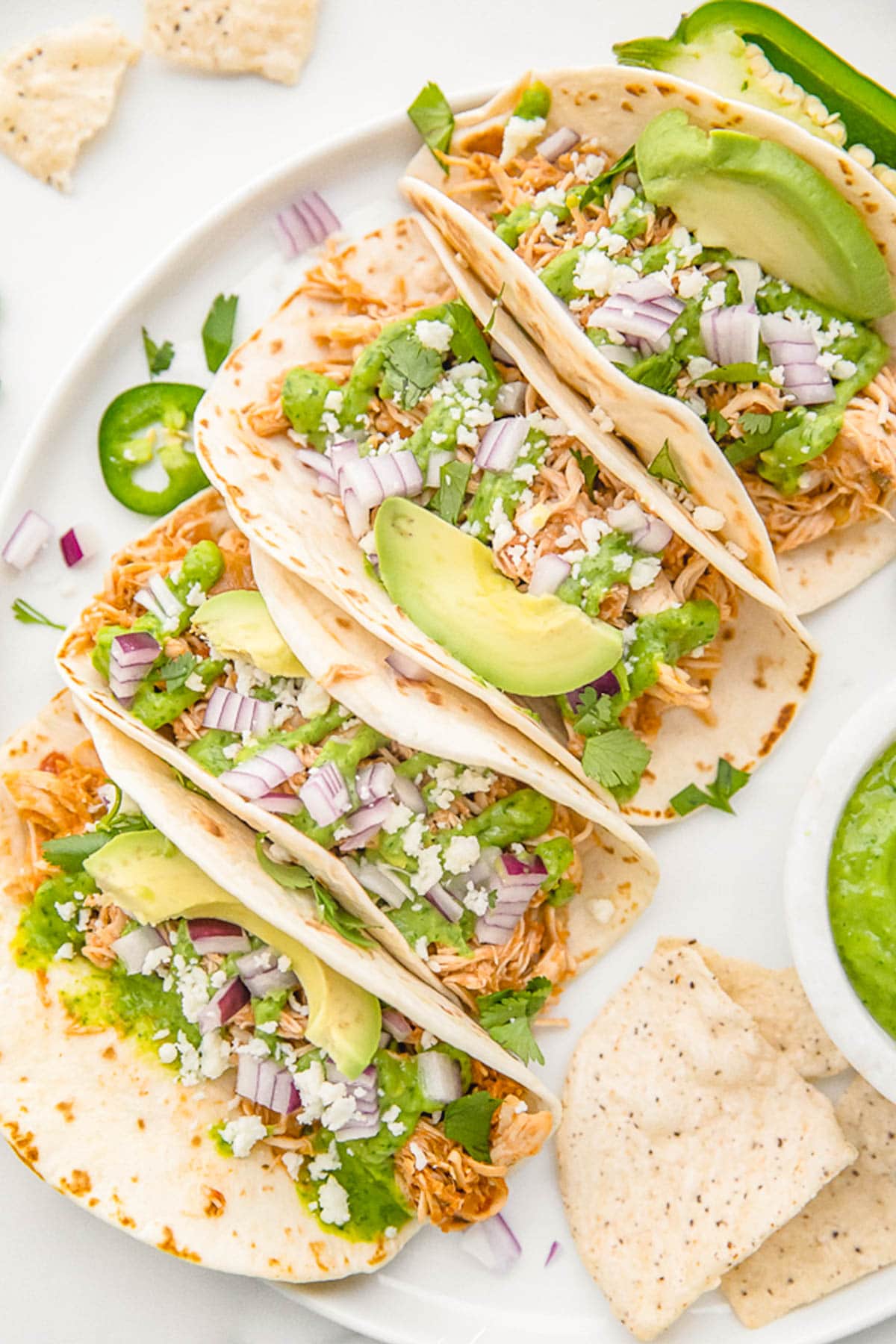 Crockpot chicken tacos arranged on a white plate. The tacos are topped with diced red onion, slices of avocado, and crumbled cheese.