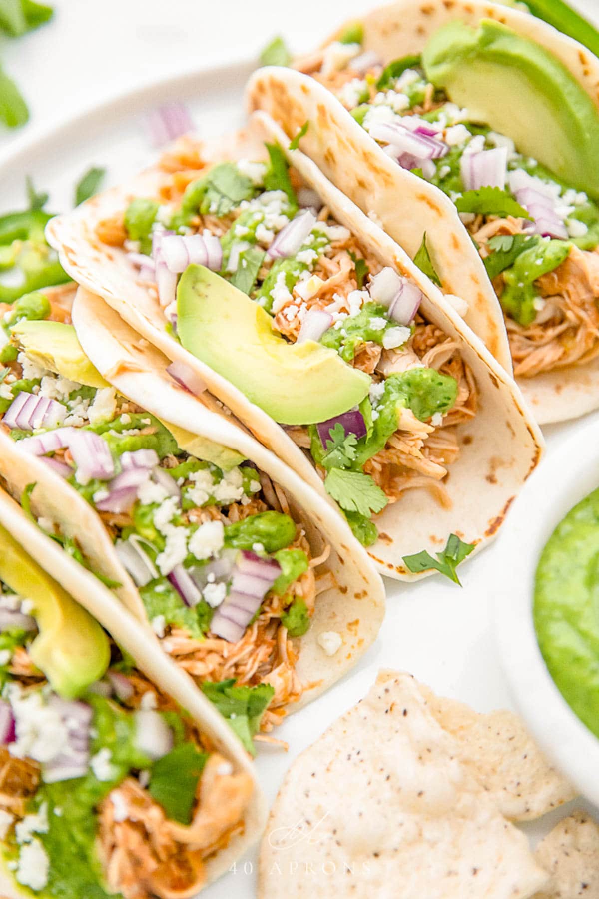 Crockpot chicken tacos arranged on a white plate. The tacos are topped with diced red onion, slices of avocado, and crumbled cheese.