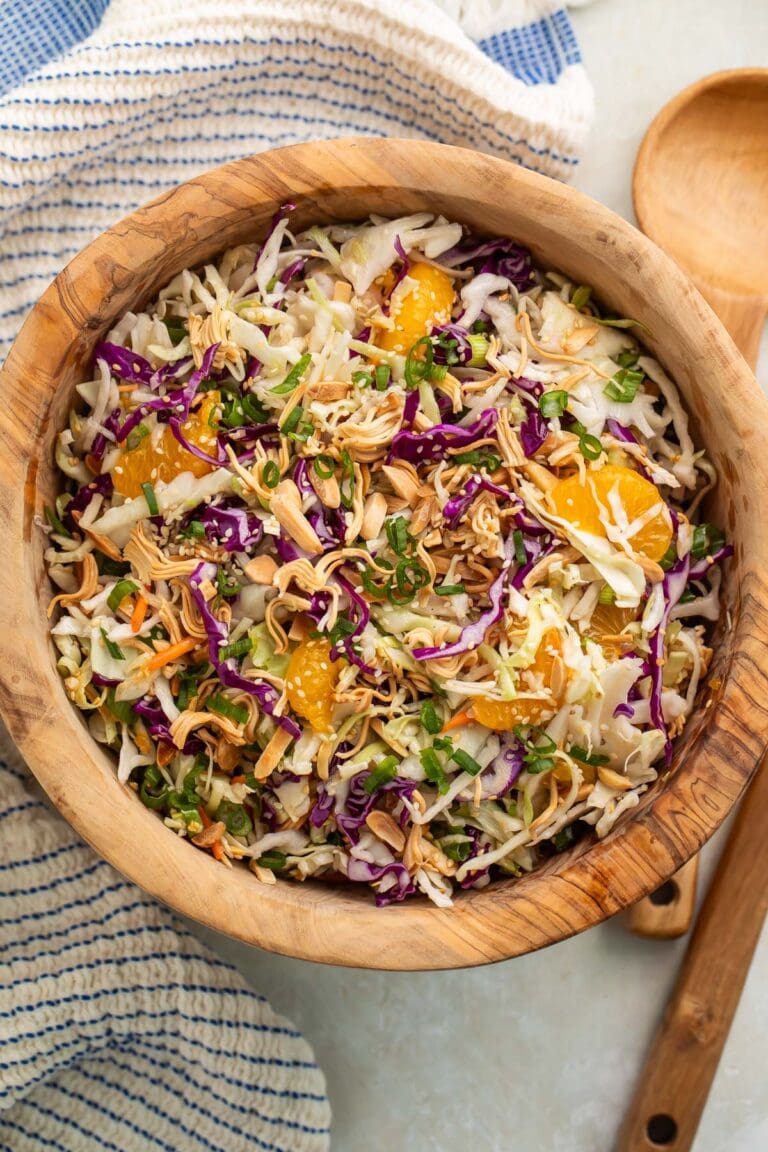 A wooden bowl of Asian slaw on a table next to a wooden spoon and a blue and white striped kitchen towel.