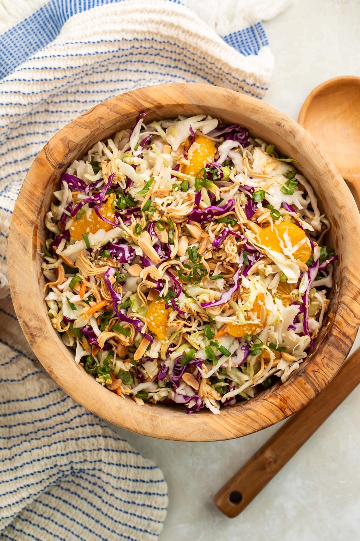 A wooden bowl of Asian slaw on a table next to a wooden spoon and a blue and white striped kitchen towel.