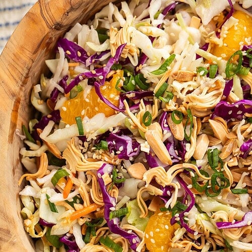 Close-up of a wooden bowl of Asian slaw on a table next to a wooden spoon and a blue and white striped kitchen towel.