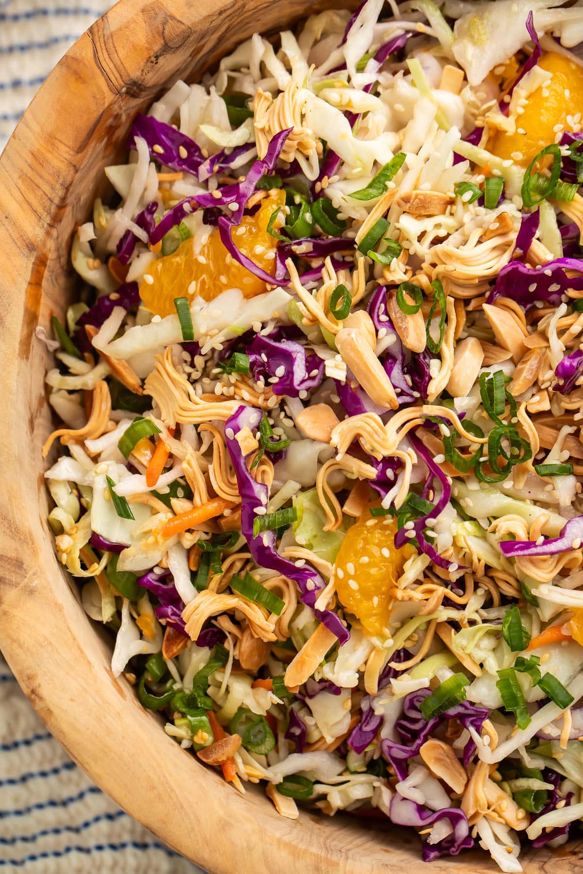 Close-up of a wooden bowl of Asian slaw on a table next to a wooden spoon and a blue and white striped kitchen towel.