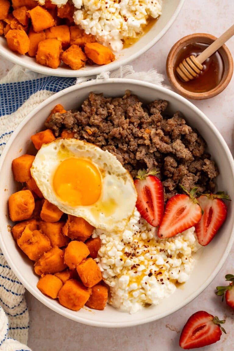 A breakfast bowl with cottage cheese, sweet potatoes, breakfast sausage, and sliced strawberries, topped with a fried egg.
