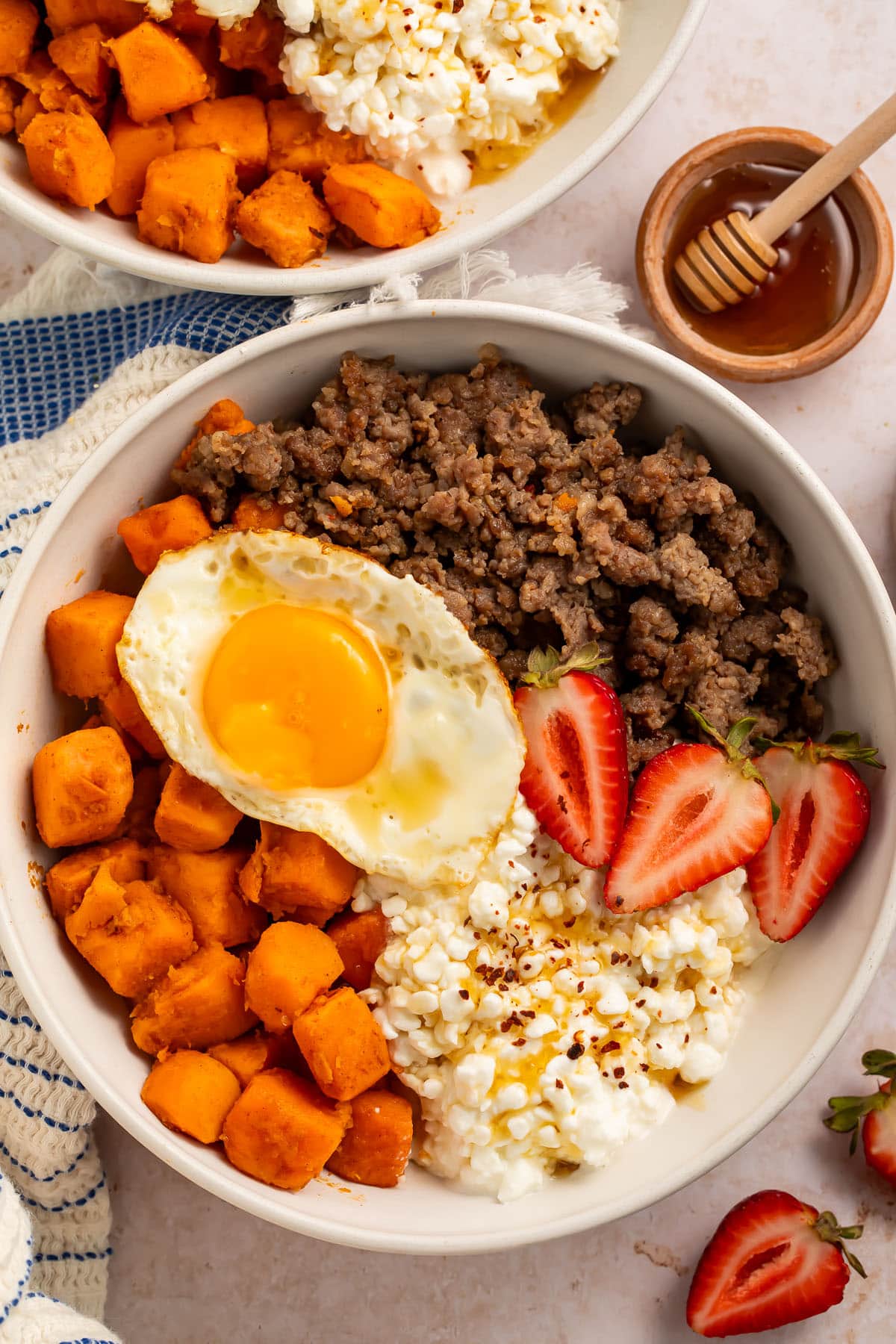 A breakfast bowl with cottage cheese, sweet potatoes, breakfast sausage, and sliced strawberries, topped with a fried egg.