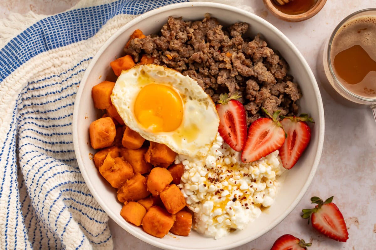 A breakfast bowl with cottage cheese, sweet potatoes, breakfast sausage, and sliced strawberries, topped with a fried egg.